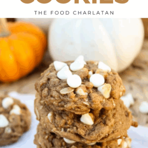 A stack of Pumpkin Oatmeal White Chocolate Chip Cookies rests on parchment paper, with white chocolate chips on top and blurred orange and white pumpkins in the background.