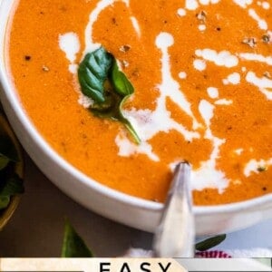 A bowl of creamy tomato soup garnished with a swirl of cream and a fresh basil leaf, with a spoon resting in the bowl. Text at the bottom reads, Easy Tomato Soup. Try this classic tomato soup recipe for a cozy meal.