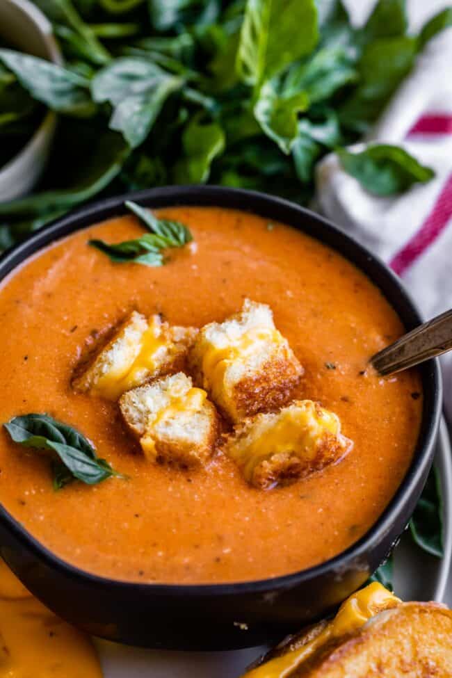 creamy tomato soup with grilled cheese floating on top with a spoon to the side.