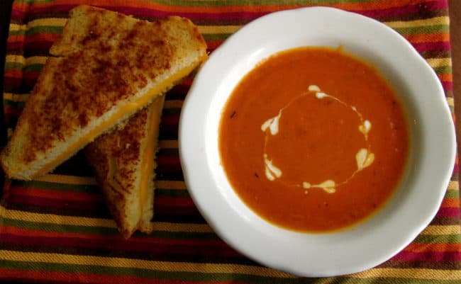 Homemade tomato soup in a bowl with heart shaped swirls of cream on top and a grilled cheese sandwich. 
