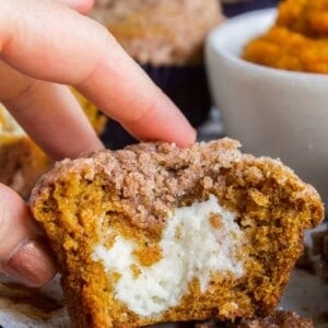 A hand holds a pumpkin cream cheese muffin cut in half, revealing a creamy white filling inside. The muffin has a crumbly topping and sits on parchment paper, surrounded by more muffins and a bowl of pumpkin puree in the background.