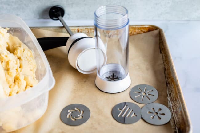 oxo plastic cookie press on a baking sheet with metal cookie plates with cookie dough on the side.