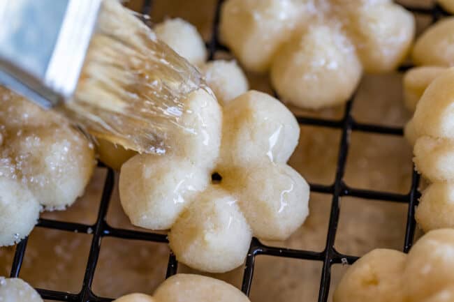 brushing corn syrup on flower shaped spritz cookies on a cooling rack.