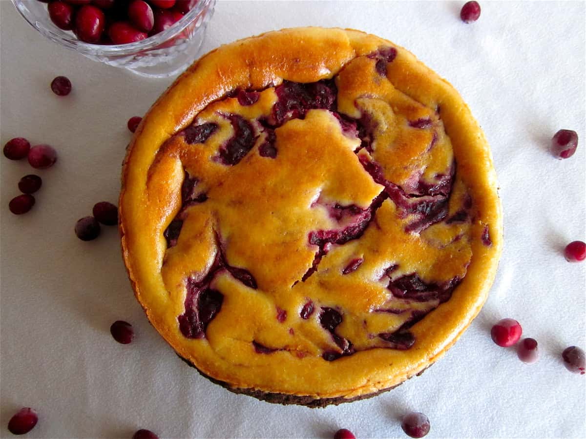 Whole cranberry cheesecake top-down view with cranberries on white tablecloth.