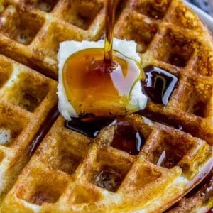A close-up of golden waffles topped with a pat of butter as maple syrup is being poured on top. Sliced strawberries and blueberries add a burst of color in the background.