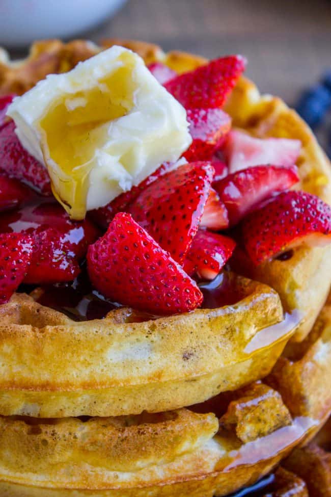 a stack of homemade waffles with whipped butter, syrup, and fresh strawberries.