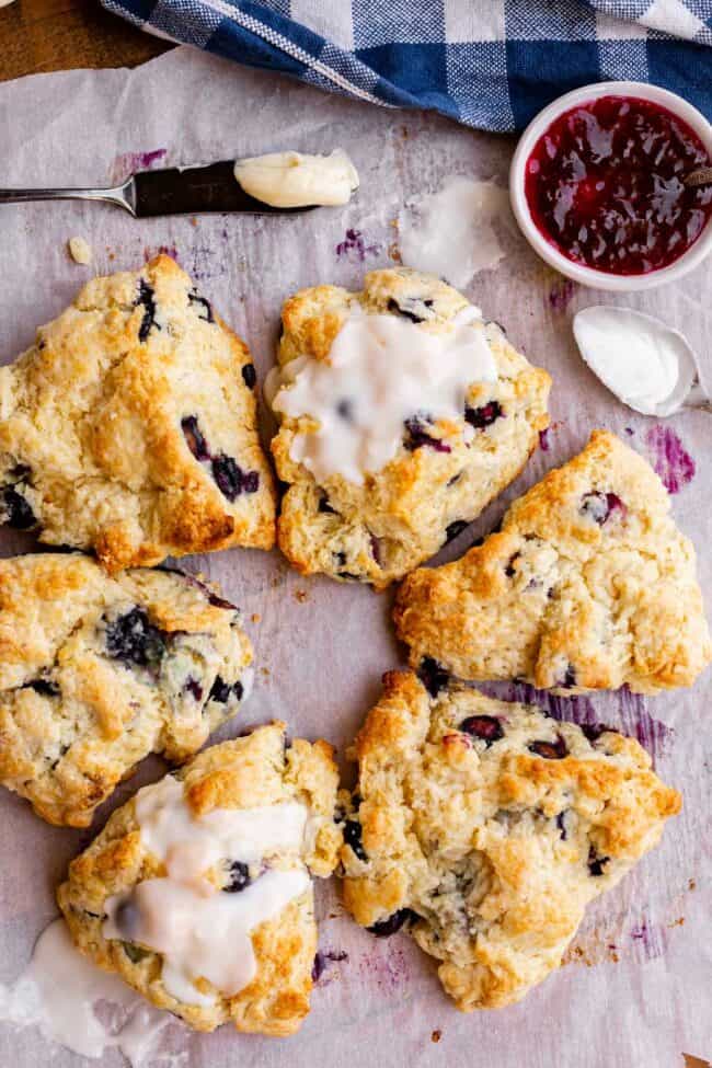 a circle of scones with glaze drizzled over some, butter and jam nearby.