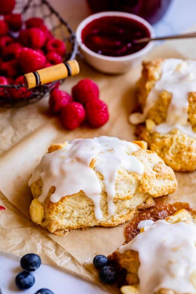 glazed scones on parchment paper with fresh raspberries and jam in the background.