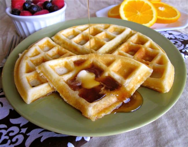homemade waffle on a plate with butter and syrup with fresh fruit in the background.