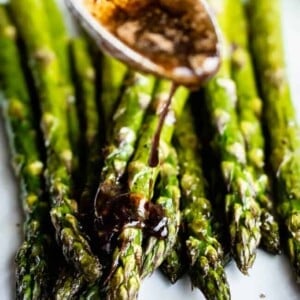 Close-up of oven roasted asparagus spears on a white plate, drizzled with a dark balsamic sauce from a spoon.