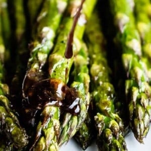 Close-up of oven roasted asparagus spears being drizzled with a dark balsamic glaze. The vibrant green asparagus tips glisten with the rich sauce.