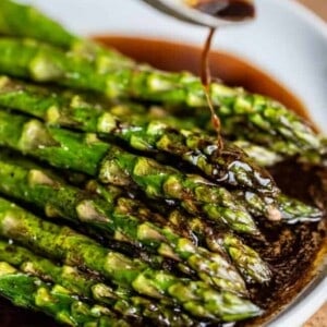 A close-up of oven roasted asparagus spears on a plate, with a dark sauce being poured over them from a spoon.