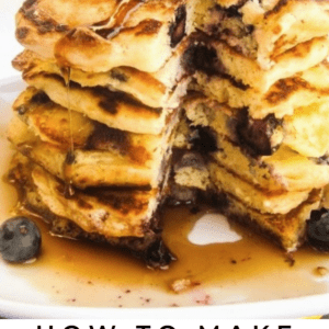 A tall stack of fluffy pancakes filled with blueberries, topped with sliced bananas, extra blueberries, and syrup, with a bite taken out. Text below reads: Lemon Blueberry Pancakes.