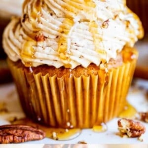 A pumpkin cupcake topped with swirled maple pecan frosting, drizzled with syrup, and garnished with a pecan. Text at the bottom reads: Pumpkin Cupcakes with Maple Pecan Frosting.