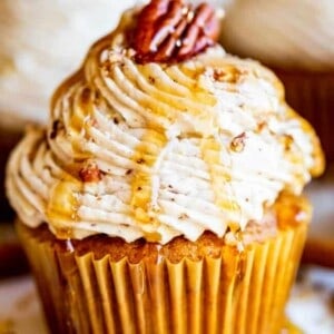 A pumpkin cupcake topped with swirled frosting, drizzled with caramel sauce, and garnished with a whole pecan. Crushed pecans and caramel are also scattered on the plate around the cupcake.
