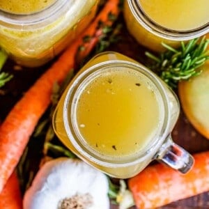 Top view of jars filled with turkey stock, surrounded by fresh carrots, garlic, herbs, and leafy greens. Text overlay reads: Turkey stock from the bones—crockpot or stovetop methods.