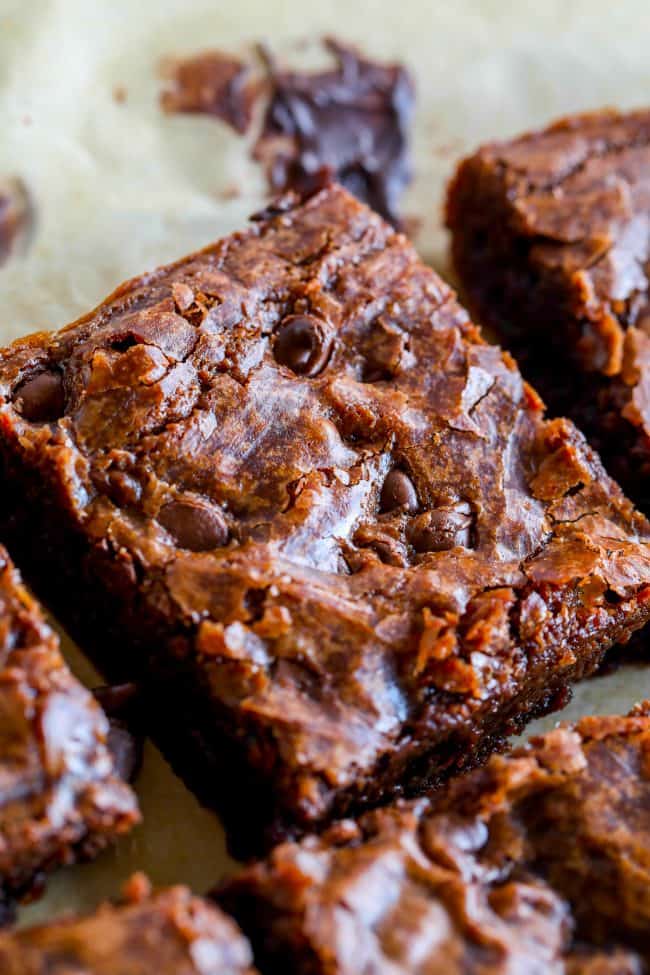 A close-up of rich, fudgy brownies with a shiny, crackly top and chocolate chips, surrounded by other brownie pieces on parchment paper.
