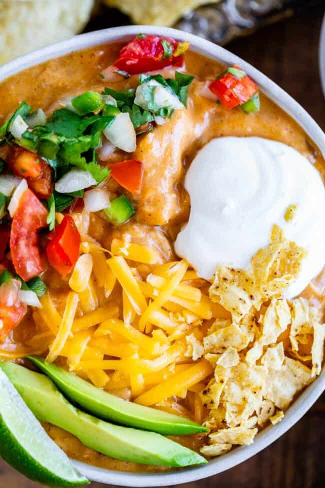 A bowl of creamy chicken enchilada soup topped with shredded cheddar cheese, sour cream, pico de gallo, crushed tortilla chips, avocado slices, and a lime wedge.