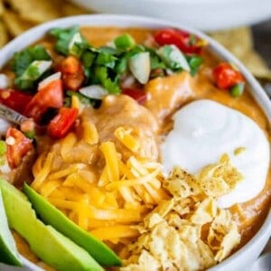 A bowl of cheesy nacho dip inspired by chicken enchilada soup, topped with shredded cheese, sour cream, chopped tomatoes, onions, cilantro, sliced avocado, and crushed tortilla chips.