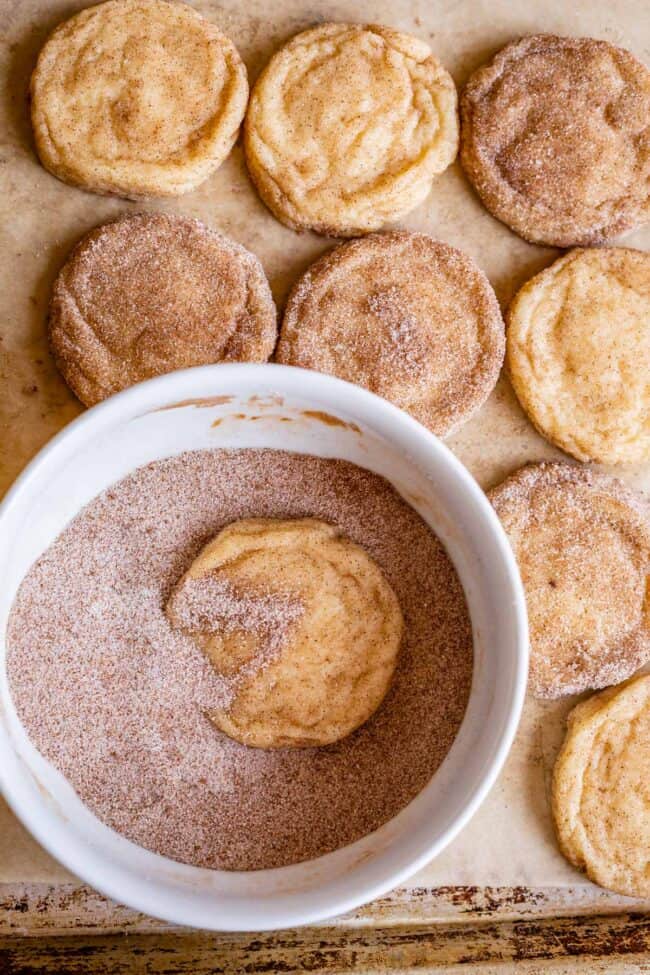snickerdoodle cookie in a bowl of cinnamon sugar, with more cookies on pan.