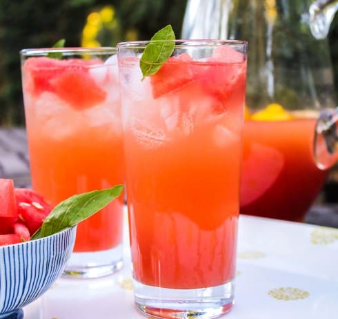 Two glasses full of watermelon lemonade with basil leafs artfully placed at the top next to a bowl of watermelon cubes.