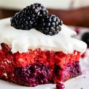 A close-up of a slice of blackberry cake with layers of pink and purple berries, topped with white frosting and two fresh blackberries, on a white plate.
