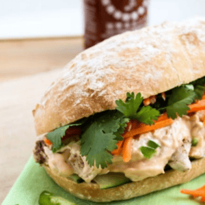 A turkey banh mi sandwich sits on a green napkin, filled with meat, cilantro, pickled carrots, and cucumbers. Sliced jalapeños are in front of the sandwich, and a bottle of sriracha sauce is in the background.