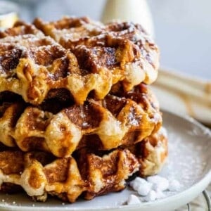 A stack of golden brown Liege waffles dusted with powdered sugar sits on a plate, with a small pitcher in the background.