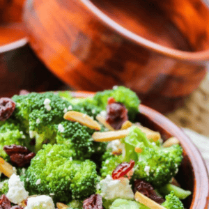 A wooden bowl filled with broccoli salad featuring cooked broccoli, dried cranberries, almond slivers, and crumbled cheese. In the background, another similar bowl is partially visible. Text above reads Broccoli Side Dish.