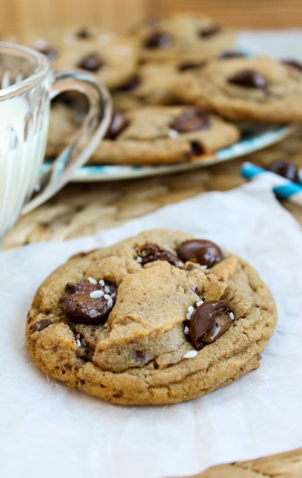 a single caramel butterscotch cookie in front of a plate full of cookies and a glass of milk. 