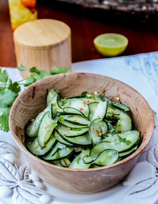 Mexican cucumber salad with cilantro and lime in a wooden salad bowl on a white platter with cilantro and lime in background. 