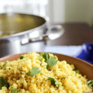 A bowl of vibrant indian cauliflower rice, garnished with fresh cilantro, sits on a table beside a pot and kitchen towel. Text above reads Indian-Spiced Cauliflower Rice.