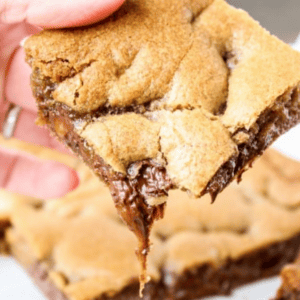 A hand holds a gooey snickers brownie square above a tray of similar brownies. The brownie is golden brown with visible caramel and chocolate filling. Text above reads, Brown Butter Caramel Brownies.