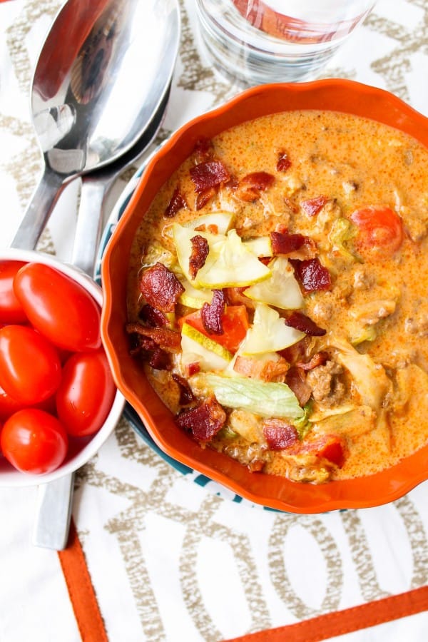 cheeseburger soup garnished with lettuce, bacon, and tomatoes in a bowl with spoons nearby.