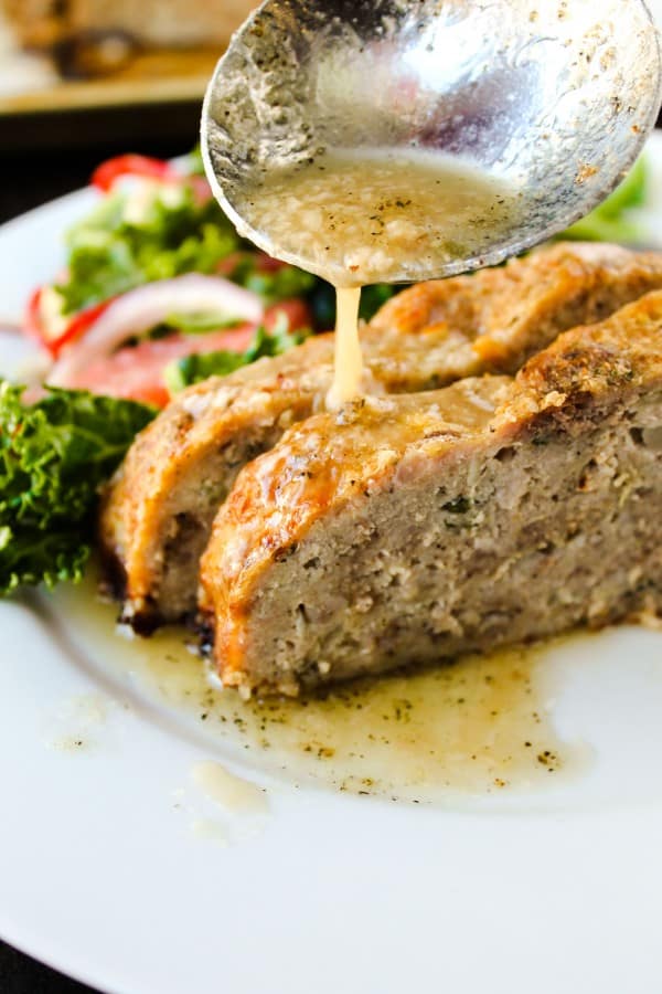 sliced meatloaf on a plate with a side salad, garlic sauce being poured onto the meatloaf.