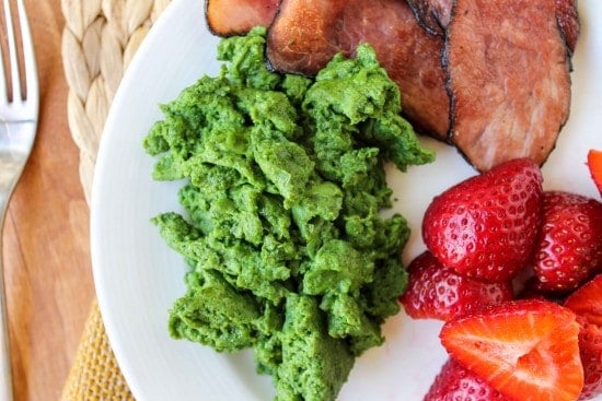 green scrambled eggs with black forest ham and strawberries on a white plate.