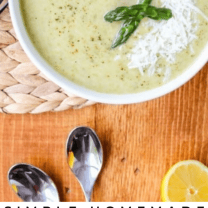 A bowl of creamy asparagus soup garnished with shredded cheese and asparagus tips sits on a woven mat. Two spoons and a lemon slice rest nearby on a wooden surface. Text reads Simple Asparagus Soup.