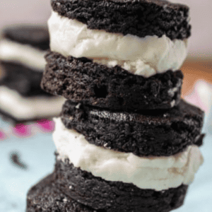 A stack of Homemade Brownie Oreos, each made with two round layers of dark chocolate brownie and a thick layer of white cream filling in the middle, arranged on blue parchment paper.