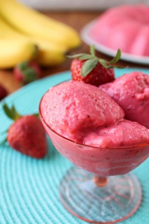 Scooped banana jello in a pink dish with bananas and strawberries in the background.