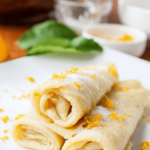 Three rolled lemon crepes are stacked on a white plate, garnished with lemon zest and powdered sugar. In the background, blurred kitchen utensils and ingredients set the scene. Text at the top reads “How To Make Lemon Crepes.”.