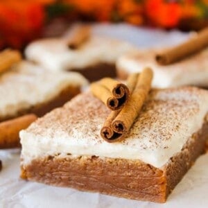 A close-up of a frosted pumpkin cake bar topped with cinnamon sticks, with more pumpkin bars and autumn leaves blurred in the background.