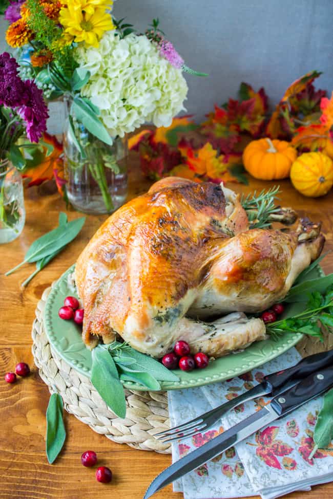 Herb roasted turkey on a platter with fall flowers and mini pumpkins in the background. 