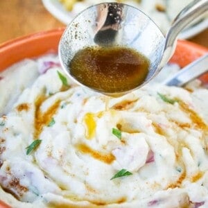 A ladle pours brown gravy over a bowl of browned butter mashed potatoes with parmesan, garnished with fresh herbs. A plate of food is blurred in the background.