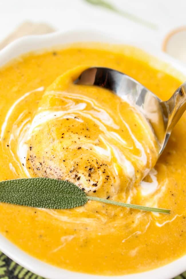 a soup spoon being dipped into a bowl of butternut squash soup with cream, fresh sage, and freshly ground pepper.