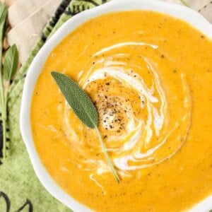 A bowl of creamy roasted butternut squash soup, swirled with cream and topped with black pepper and a fresh sage leaf, sits on a green and beige patterned cloth with extra sage leaves nearby.