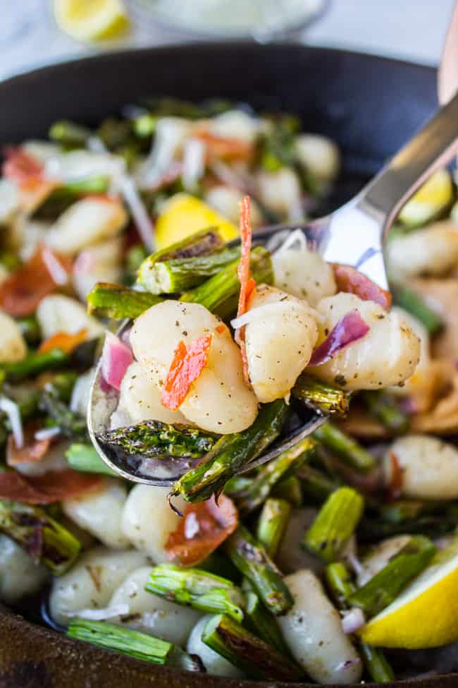 a metal spoon lifting asparagus, prosciutto, and gnocchi from a pan.