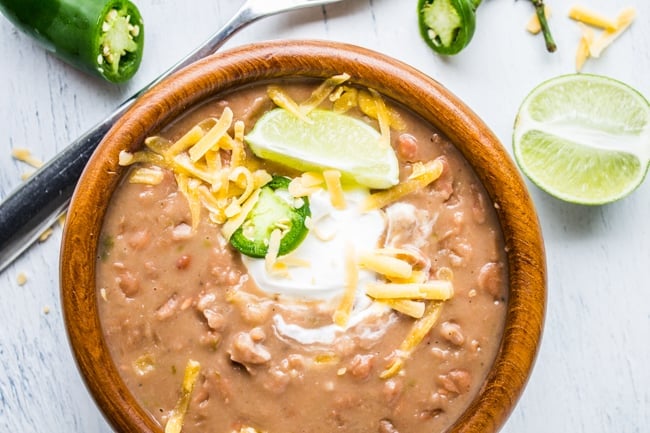 overhead shot of mexican pinto beans in a wooden bowl topped with cheese, jalapeño, sour cream, and lime.