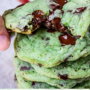 A hand holds a soft, green Mint Chocolate Chip Cookie with melted chocolate above a stack of similar cookies. Text at the bottom reads: Soft & Chewy Mint Chocolate Chip Cookies.