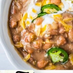 A bowl of Mexican beans topped with melted cheese, sour cream, sliced jalapeños, and a lime wedge. The image includes the text Tejano Pinto Beans at the bottom.
