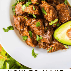 A white plate with Tejano Carne Guisada, garnished with fresh cilantro and a slice of avocado. Text below reads: Tejano Carne Guisada – The Food Charlatan.